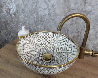 May include: A patterned ceramic sink with a black and white design and a gold trim. A brass tap is positioned above the sink, with water flowing into the basin. The sink is set on a wooden surface.