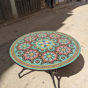 Peut inclure: Une table ronde dotée d'un plateau en mosaïque présentant un motif géométrique complexe dans des tons turquoise, rouge, jaune, noir et blanc. La table est soutenue par un cadre en métal noir avec quatre pieds incurvés partant du centre.