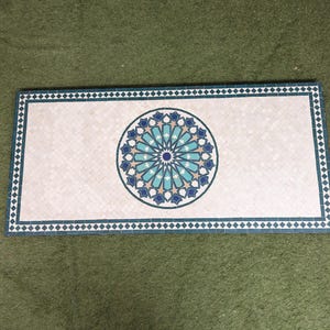 May include: A white ceramic tile with a blue and green geometric pattern in the center. The tile is surrounded by a blue and white border.