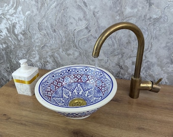 Può includere: Un lavabo decorativo in ceramica con un motivo floreale blu e bianco. Un rubinetto in ottone è posizionato sopra il lavabo, con acqua che scorre nel bacino. A sinistra c'è un dispenser di sapone bianco con dettagli dorati.