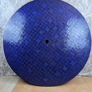 May include: A circular table top with a mosaic tile design in various shades of blue. The surface is patterned with small, square and hexagonal tiles, and has a central hole. The tiles create a textured, reflective surface.