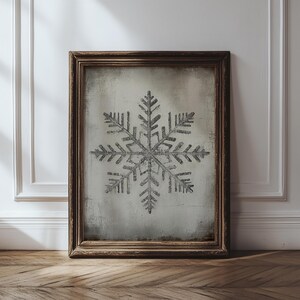 Puede incluir: Impresión enmarcada con un diseño de copo de nieve en tonos grises. La obra de arte se encuentra sobre un fondo texturizado blanquecino y está enmarcada en madera marrón oscuro. La impresión tiene un tema invernal.