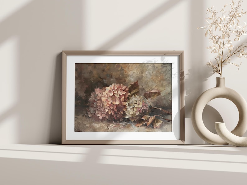 May include: A framed floral print featuring a cluster of pink and white hydrangeas with brown leaves. The artwork is displayed on a white surface, with a beige vase and decorative branch in the background.
