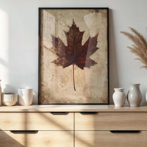 May include: A framed print featuring a large, detailed maple leaf in shades of brown and red against a textured, beige background. The artwork is displayed on a wooden dresser, alongside white vases and decorative plants.
