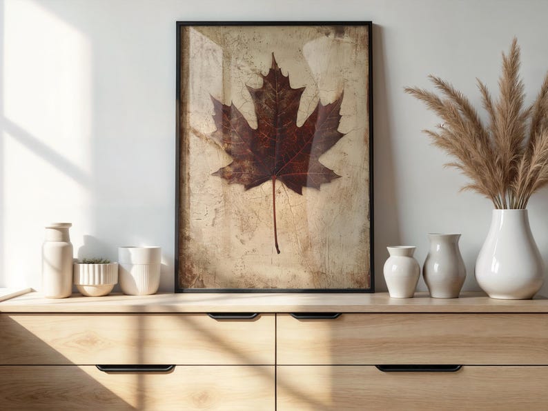 May include: A framed print featuring a large, detailed maple leaf in shades of brown and red against a textured, beige background. The artwork is displayed on a wooden dresser, alongside white vases and decorative plants.