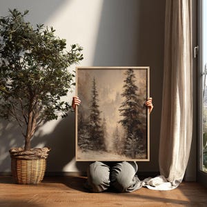 May include: A framed landscape painting of a misty forest with tall evergreen trees. The artwork is held by a person, with the bottom half of their body visible. The painting has a neutral colour palette and is displayed indoors.
