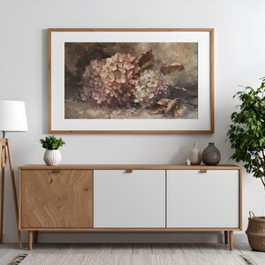 May include: A framed floral print featuring a cluster of pink and white hydrangeas. The artwork is displayed above a wooden cabinet with white doors. A floor lamp and potted plant are also in the room.