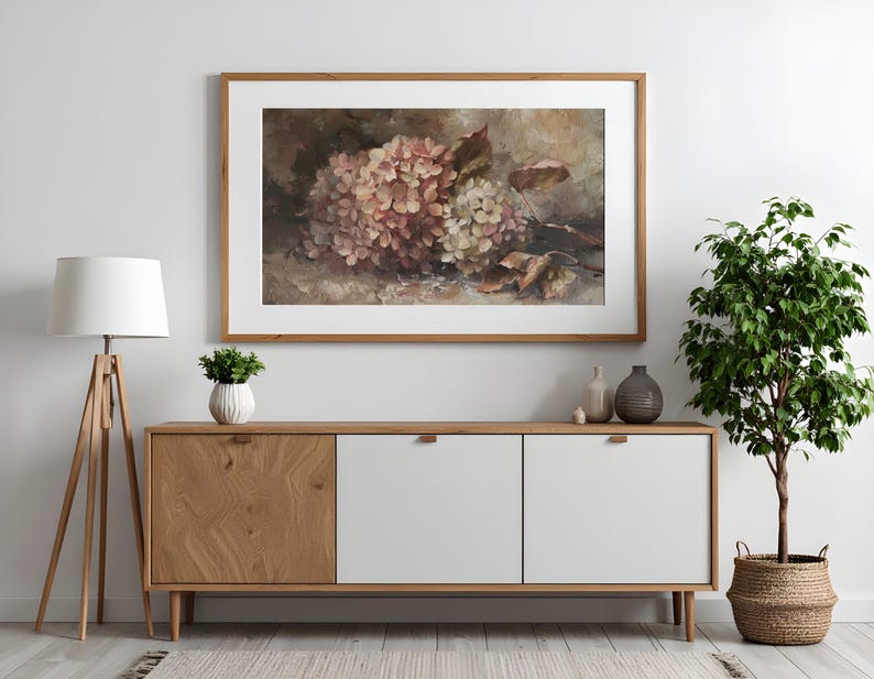 May include: A framed floral print featuring a cluster of pink and white hydrangeas. The artwork is displayed above a wooden cabinet with white doors. A floor lamp and potted plant are also in the room.