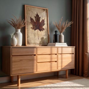 May include: A light-colored wooden sideboard with three drawers and two doors. The sideboard is topped with a framed leaf print, vases, and books. The room has a neutral color palette with a window and curtains.