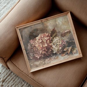 May include: A framed floral print featuring a cluster of pink and cream hydrangeas. The artwork is set in a light wooden frame and rests on a brown upholstered surface. The painting has a vintage aesthetic.