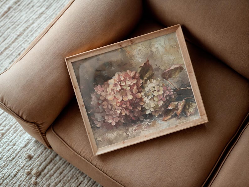 May include: A framed floral print featuring a cluster of pink and cream hydrangeas. The artwork is set in a light wooden frame and rests on a brown upholstered surface. The painting has a vintage aesthetic.