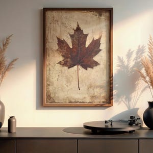 May include: A framed print featuring a large, detailed brown maple leaf against a textured background. The artwork hangs above a dark gray cabinet, with decorative vases and a record player. The overall theme is nature and home decor.