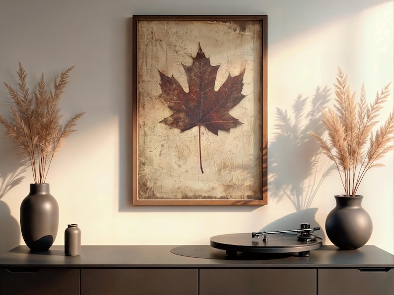 May include: A framed print featuring a large, detailed brown maple leaf against a textured background. The artwork hangs above a dark gray cabinet, with decorative vases and a record player. The overall theme is nature and home decor.