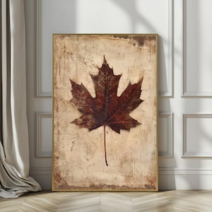 May include: A framed print featuring a detailed, dark brown maple leaf against a textured, beige background. The leaf's veins are visible, and the frame is gold. The artwork evokes a sense of autumn and nature.