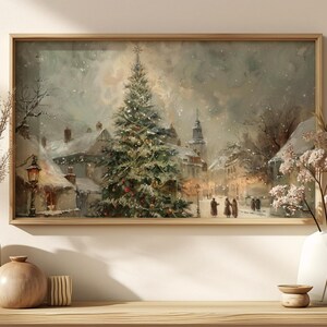 Peut inclure: Peinture encadr&eacute;e d'un paysage hivernal repr&eacute;sentant une place de ville enneig&eacute;e avec un sapin de No&euml;l d&eacute;cor&eacute;. L'&oelig;uvre pr&eacute;sente un clocher d'&eacute;glise, des lampadaires et des personnages marchant dans la neige, le tout dans des tons sourds.