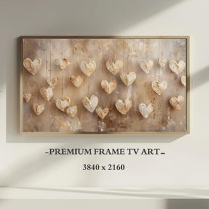 Puede incluir: Una pieza de arte mural rectangular en un marco dorado con un fondo abstracto beige y crema con múltiples formas de corazón texturizadas y tridimensionales que cuelgan de finas líneas verticales. El texto dice PREMIUM FRAME TV ART 3840 x 2160.