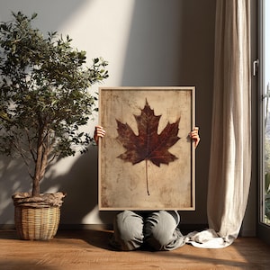 May include: A framed print featuring a large, detailed maple leaf in shades of brown and red. The leaf is set against a textured, beige background. The artwork is held by a person in a room with a tree in a woven basket and a curtain.