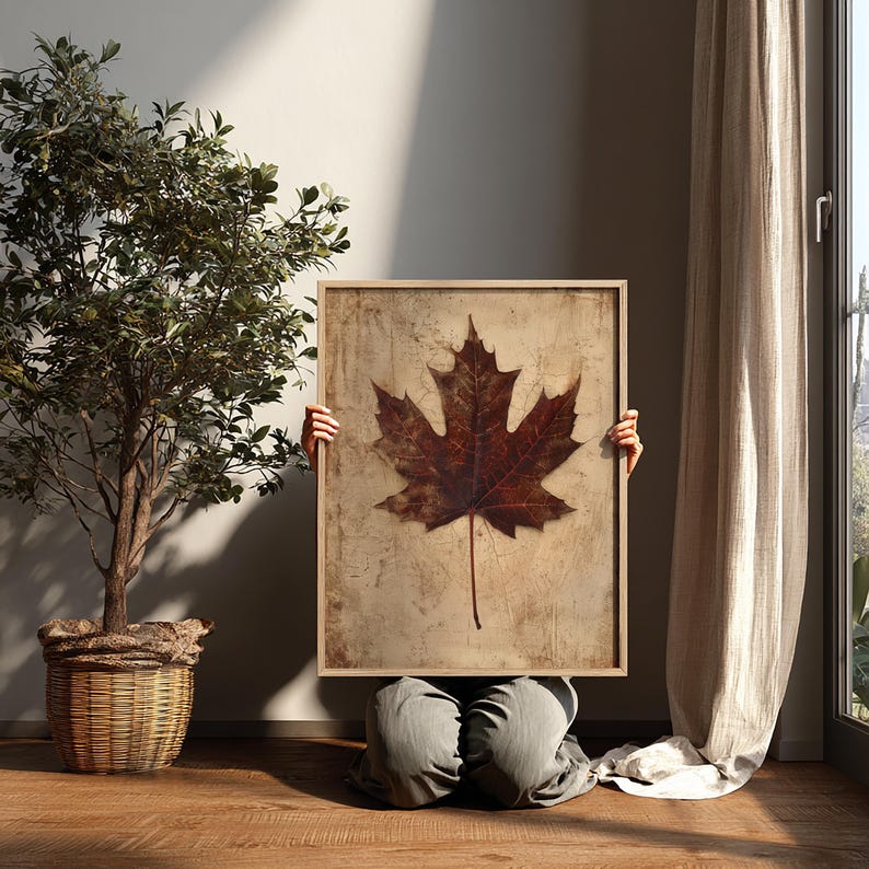May include: A framed print featuring a large, detailed maple leaf in shades of brown and red. The leaf is set against a textured, beige background. The artwork is held by a person in a room with a tree in a woven basket and a curtain.