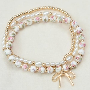 May include: Three-strand bracelet set. The top strand is gold-colored beads, the middle strand has white floral beads and pearls, and the bottom strand has white floral beads, pearls, and a gold-colored bow charm.