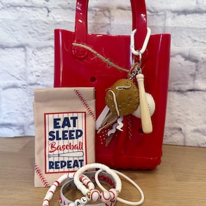 May include: A red tote bag with a baseball-themed keychain, a small pouch with "EAT SLEEP BASEBALL REPEAT" text, and several white and red baseball bracelets. The keychain features a baseball glove and bat.