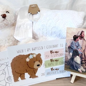Könnte beinhalten: Eine Sammlung religiöser Artikel, darunter ein weißer Teddybär, eine weiße Plüschdecke, eine Grußkarte mit einer Bärenillustration und Text sowie ein gerahmtes Bild von Jesus mit Kindern. Die Karte lautet: "Wenn ich getauft bin, verpflichte ich mich zu: Meinem Zeugnis..."