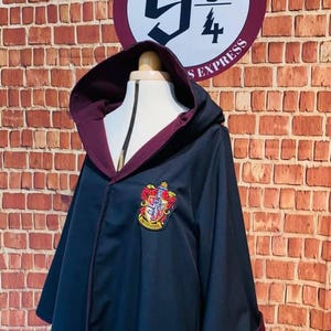Puede incluir: Una túnica negra con capucha y forro burdeos y un escudo de Gryffindor bordado en la parte delantera. La túnica se muestra contra una pared de ladrillo con un cartel que dice "Platform 9 3/4" y "Express".
