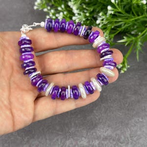 May include: A beaded bracelet featuring a mix of purple and white beads. The bracelet has a silver clasp and is held in a hand. The beads are a mix of dark and light purple, with some white beads interspersed. The bracelet is a stylish accessory.