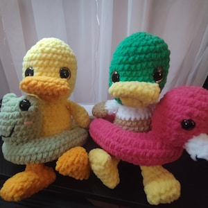 May include: Two crocheted duck plushies, one yellow with a green frog on its back and one green with a pink flamingo on its back. Both ducks are wearing crocheted life preservers.