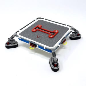 Puede incluir: Una plataforma cuadrada gris y blanca con un diseño rectangular rojo en el centro. La plataforma tiene cuatro patas ajustables con bases negras. La plataforma es azul en los lados y tiene detalles amarillos.