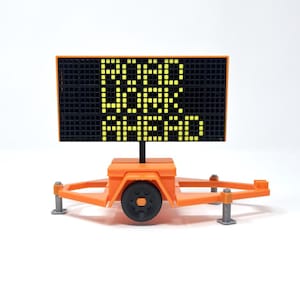 Può includere: Un cartello arancione di lavori stradali su un rimorchio, che mostra il testo "ROAD WORK AHEAD" in pixel gialli illuminati. Il cartello è posto su uno sfondo bianco.