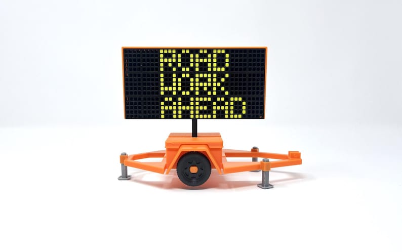 Pu&ograve; includere: Un cartello arancione di lavori stradali su un rimorchio, che mostra il testo "ROAD WORK AHEAD" in pixel gialli illuminati. Il cartello &egrave; posto su uno sfondo bianco.