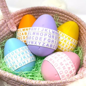 May include: A pink woven basket containing five brightly coloured Easter eggs. The eggs are decorated with a white band featuring the alphabet. The basket is lined with green paper grass. The eggs are in shades of blue, orange, purple, yellow, and pink.