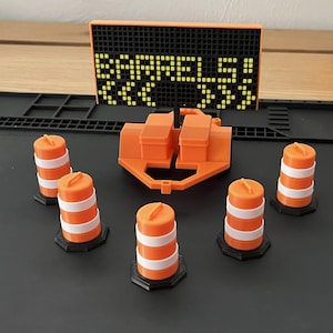 Pu&ograve; includere: Un set di costruzione stradale in miniatura arancione e bianco. Il set include coni stradali arancioni con strisce bianche, un rimorchio e un cartello con la scritta "BARRELS!". Il cartello &egrave; nero con lettere pixelate gialle.