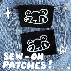 May include: Two black sew-on patches with a white frog design are displayed on a denim pocket. The patches are rectangular and feature a simple, cartoon-style frog illustration. The text "SEW-ON PATCHES!" is visible.