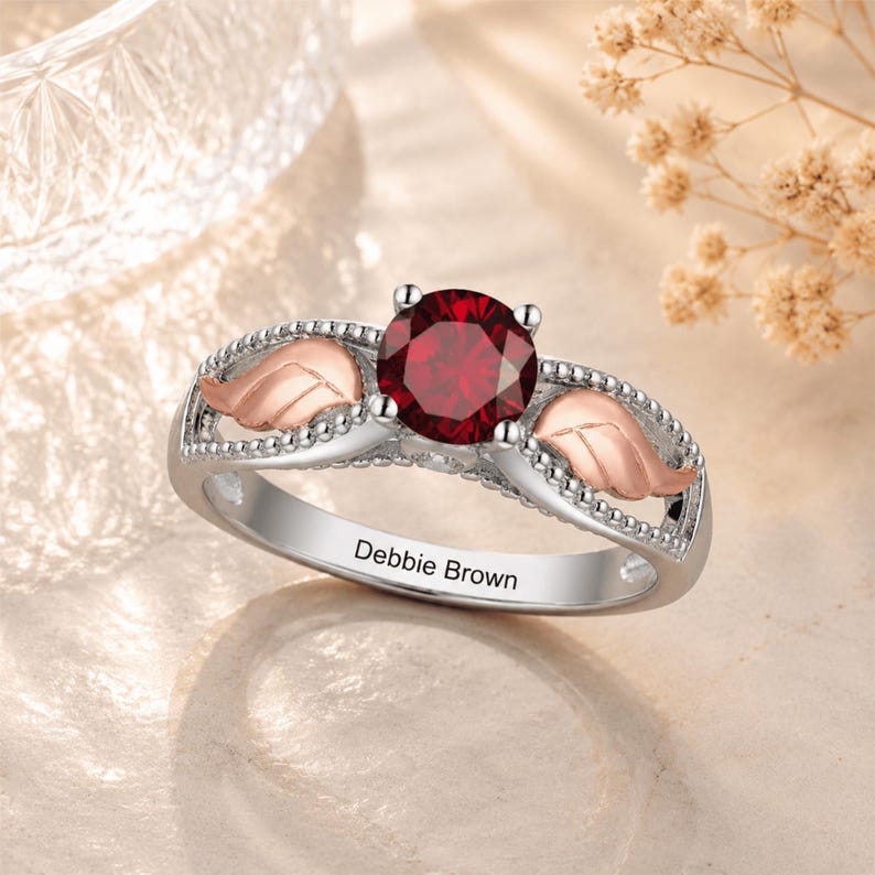 May include: A silver ring with a large, round, red gemstone as the centerpiece. The ring features rose gold leaf accents on either side of the gemstone and the inscription "Debbie Brown" on the band.