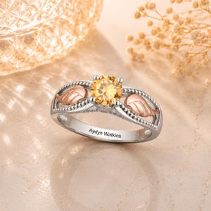 May include: A silver ring with a large, round, yellow gemstone at the center. The ring features two rose gold leaf accents and the engraved name "Aydyn Watkins". The ring is set against a soft, neutral background.