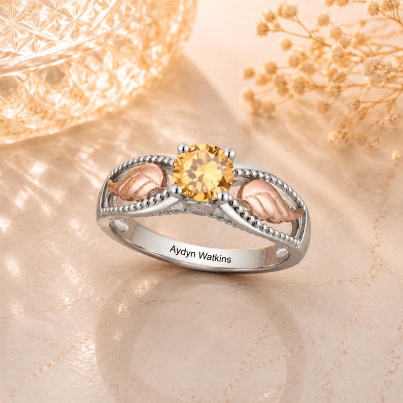 May include: A silver ring with a large, round, yellow gemstone at the center. The ring features two rose gold leaf accents and the engraved name "Aydyn Watkins". The ring is set against a soft, neutral background.
