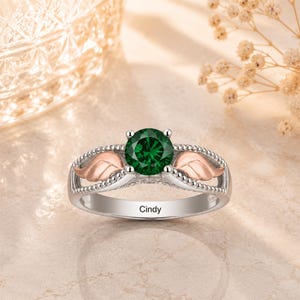 May include: A silver ring with a large, round, emerald-green gemstone. The ring features rose-gold-colored wing accents on either side of the gemstone and the name "Cindy" engraved on the band.
