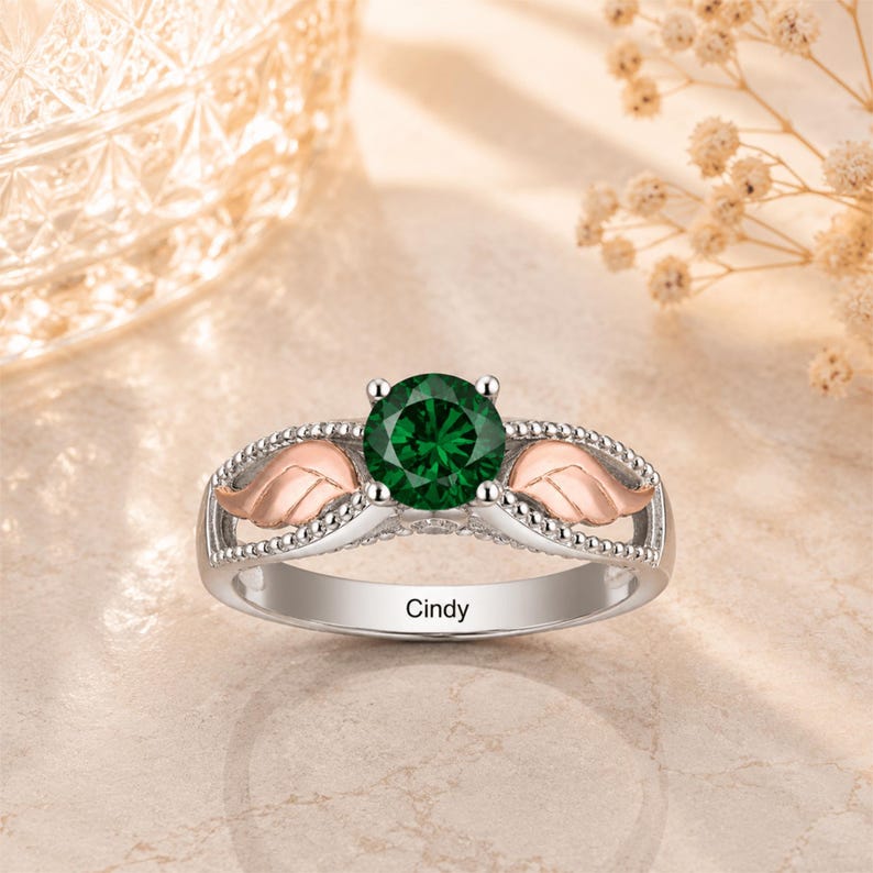 May include: A silver ring with a large, round, emerald-green gemstone. The ring features rose-gold-colored wing accents on either side of the gemstone and the name "Cindy" engraved on the band.