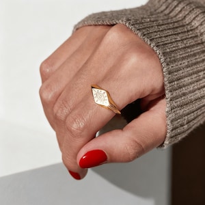 May include: A gold ring with a diamond-shaped face. The face of the ring is white with a detailed design. The ring is on a finger with red nail polish. A brown sweater is in the background.