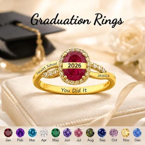 May include: A gold ring featuring a central oval red stone with the text 2026, surrounded by small clear stones. The band is engraved with Hewitt School, Jessica, and You Did It. Below are twelve circular birthstones labeled by month.