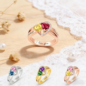 May include: Rose gold ring with two heart-shaped gemstones, one yellow and one red, surrounded by small clear stones. The ring is engraved with "Jackson" and "Rachel" on the sides and "I love you" on the band. Additional rings with different colored gemstones are also shown.