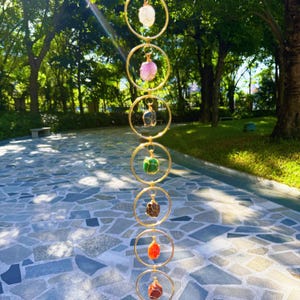 May include: A vertical hanging decoration featuring seven gemstones in various colors, suspended within gold-colored metal rings. The gemstones include white, pink, blue, green, brown, orange, and red. The rings are connected by smaller gold-colored links.