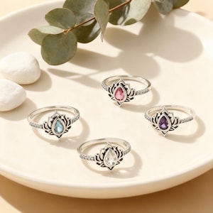 May include: Four silver rings, each with a teardrop-shaped gemstone in pink, blue, clear, and purple. The rings have a lotus flower design and a band with small decorative details. The rings are presented on a white plate with decorative elements.