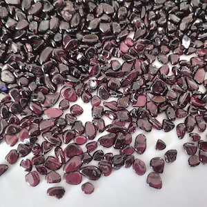 Könnte beinhalten: Nahaufnahme von zahlreichen polierten, tiefrot-violetten Edelsteinen. Die Steine sind von unterschiedlicher Größe und Form, mit einer glatten, reflektierenden Oberfläche. Die Edelsteine sind auf einer weißen Oberfläche verstreut. Ideal für Schmuckherstellung oder Dekorationszwecke.