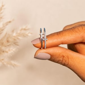 May include: A silver ring featuring a thin band and a vertical cross-shaped centerpiece. The cross is embellished with a series of small, clear, round-cut stones set along its length, providing a reflective and metallic appearance to the jewelry piece.
