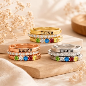 May include: Three personalised rings in gold, rose gold, and silver. Each ring features a band with a name or word, such as "Love", "Emma", and "mama". The rings are adorned with colourful gemstones and clear crystals.