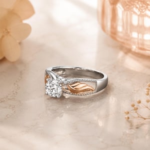 May include: A silver and rose gold engagement ring with a large, round, clear gemstone. The ring features a decorative leaf design on the sides and a polished finish. The ring is set on a light-colored surface.