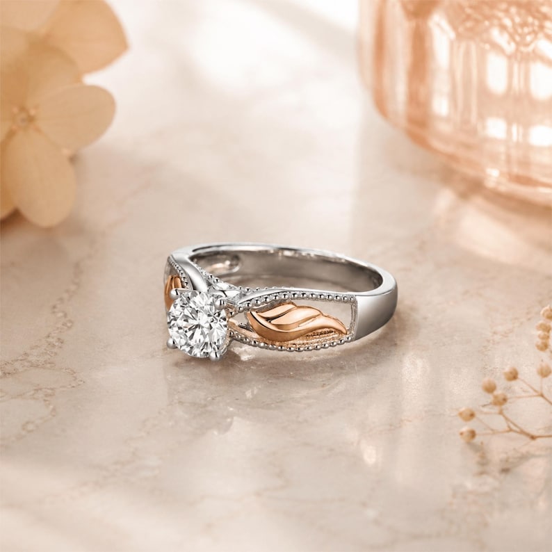 May include: A silver and rose gold engagement ring with a large, round, clear gemstone. The ring features a decorative leaf design on the sides and a polished finish. The ring is set on a light-colored surface.