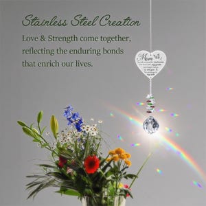 May include: A heart-shaped memorial ornament with the word "Mom" and a butterfly design, hanging from a chain. The ornament is suspended below a string of colourful beads and a clear crystal ball. A bouquet of flowers is in the foreground.